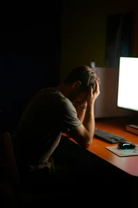 Une personne, un manager ou un dirigeant ou un salarié, stressée professionnellement qui se tient assise devant son écran d'ordinateur tard le soir. Elle se tient la tête entre les mains.